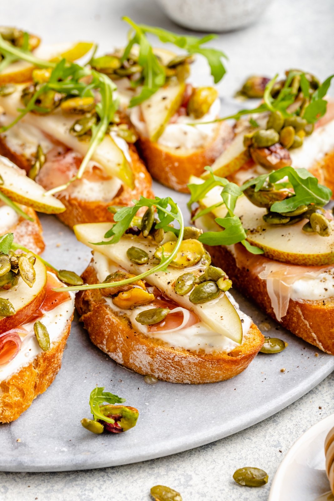 Precioso crostini de ricotta y pera con prosciutto salado, rúcula fresca y mezcla de galletas saladas de pistacho y semillas de calabaza. ¡Este simple aperitivo es la combinación perfecta de dulce, salado, cremoso y crujiente, que está garantizado para impresionar a los invitados! Espolvoree miel y un poco de sal marina encima para obtener la mejor combinación de sabores. Crostini de ricotta y pera, con nueces y rúcula, en un plato
