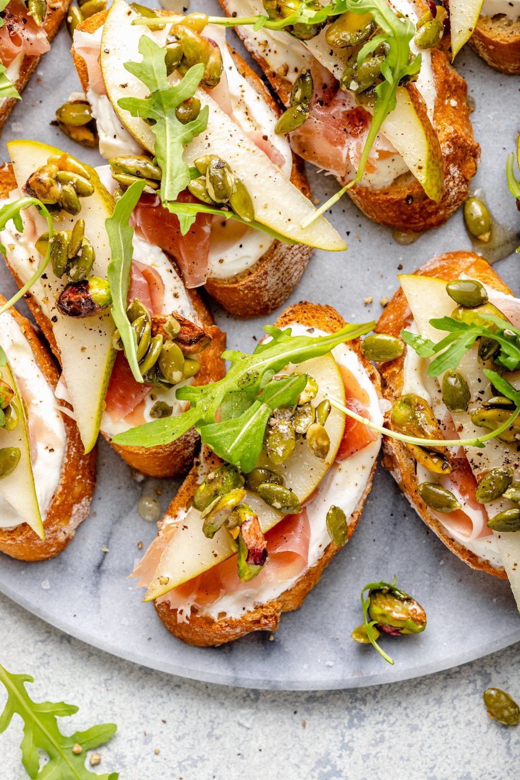 Precioso crostini de ricotta y pera con prosciutto salado, rúcula fresca y mezcla de galletas saladas de pistacho y semillas de calabaza. ¡Este simple aperitivo es la combinación perfecta de dulce, salado, cremoso y crujiente, que está garantizado para impresionar a los invitados! Espolvoree miel y un poco de sal marina encima para obtener la mejor combinación de sabores. Peras y queso ricotta con nueces y jamón en un plato
