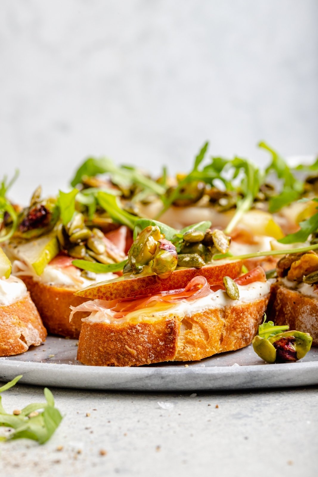 Precioso crostini de queso ricotta y pera con prosciutto salado, rúcula fresca y sabrosa mezcla de manteca de pistacho con semillas de calabaza. Este simple aperitivo es dulce, salado, cremoso y crujiente. ¡La combinación perfecta, garantizada para impresionar a los invitados! Agregue miel y espolvoree con Sal de Shanghai en la parte superior para obtener la mejor combinación de sabores. Peras simples y queso ricotta en el plato
