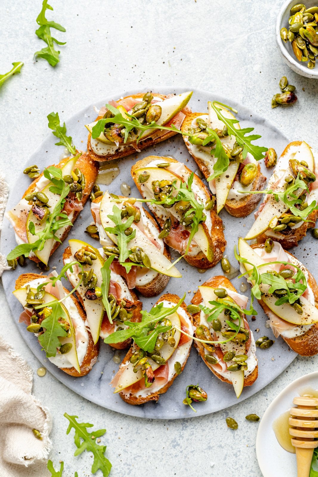 Precioso crostini de ricotta y pera con prosciutto salado, rúcula fresca y mezcla de galletas saladas de pistacho y semillas de calabaza. ¡Este simple aperitivo es la combinación perfecta de dulce, salado, cremoso y crujiente, que está garantizado para impresionar a los invitados! Espolvoree miel y un poco de sal marina encima para obtener la mejor combinación de sabores. Crostini de ricotta y pera con mezcla de rúcula y nueces en un plato