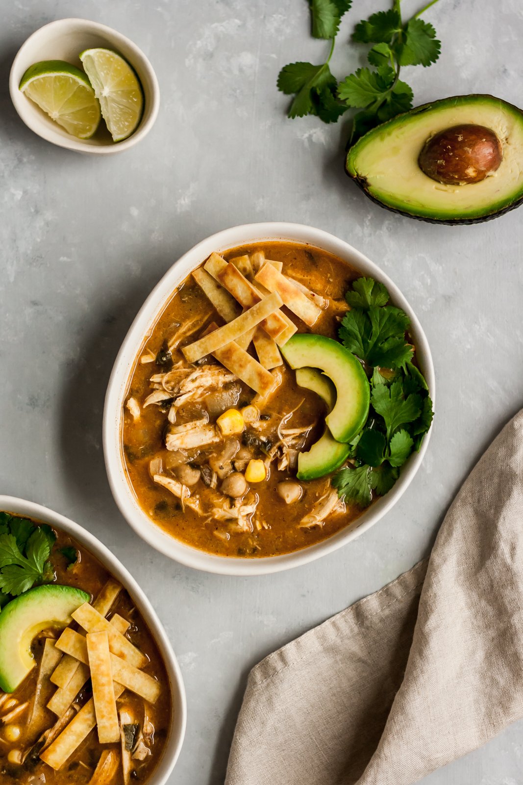 Chili de pollo blanco saludable que es fácil y sin lácteos. Elaborado con chile verde, pollo, choclo y garbanzos mixtos para que quede cremoso. ¡Una nueva receta favorita de la familia! Sirva con aguacate, totopos y cilantro. Chile de pollo blanco saludable en un tazón blanco junto a un plato de gajos de lima y medio aguacate