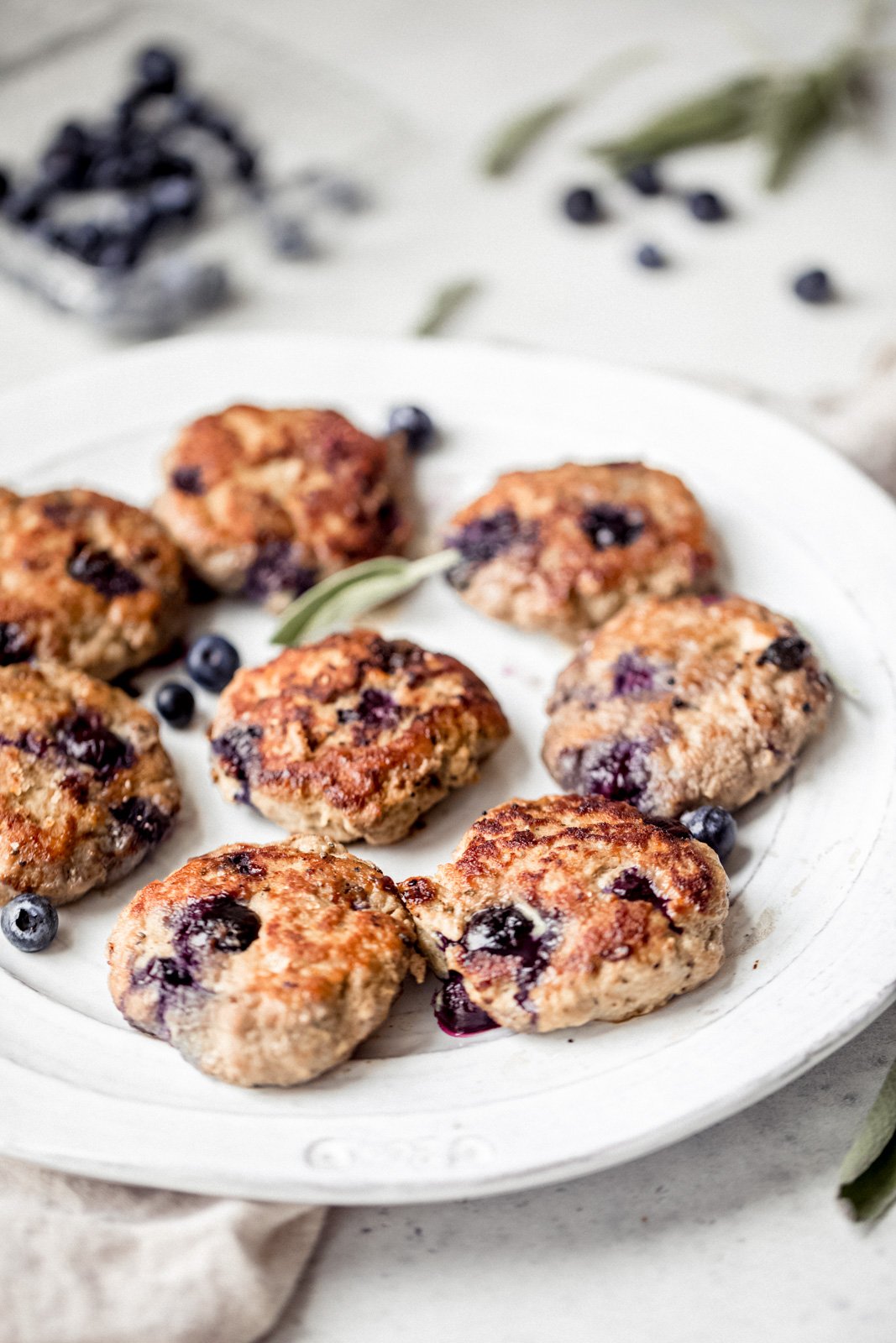 Dulces y deliciosos pays de desayuno de salchicha de pavo y arándanos Paleo Maple con deliciosas especias y jugosos arándanos en cada bocado. Sirve estas increíbles hamburguesas de salchicha de pavo con huevos fritos. ¡Disfruta del mejor desayuno rico en proteínas y bajo en carbohidratos! Pastel de salchicha de pavo en un plato con arándanos en el fondo