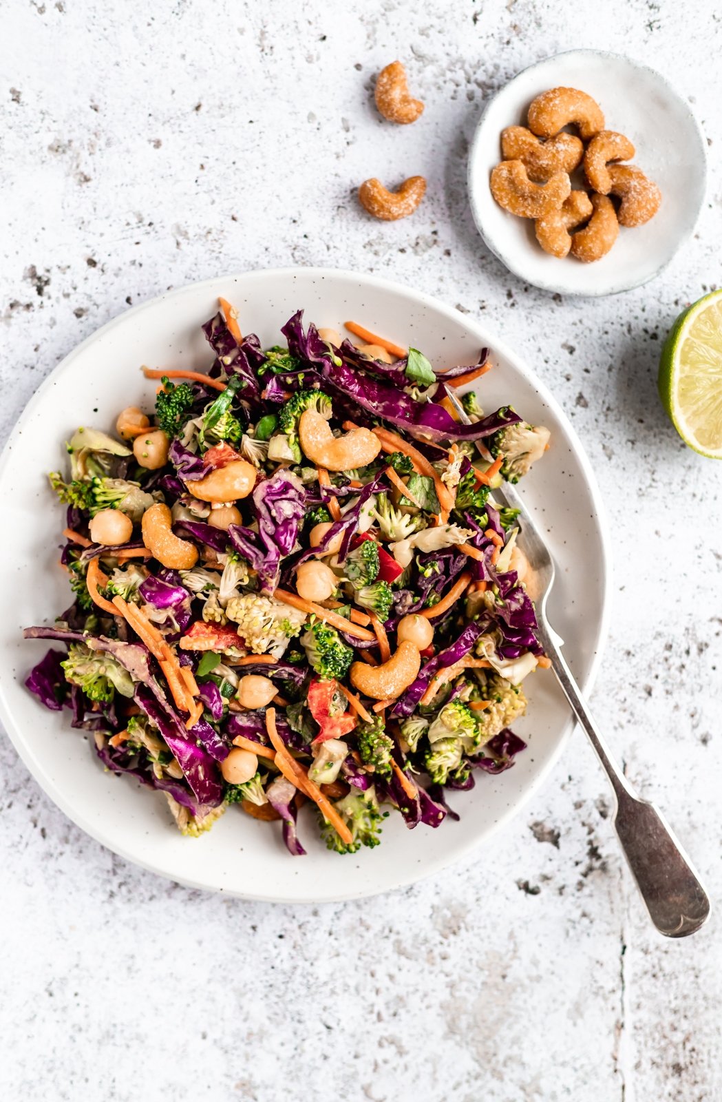 Deliciosa y saludable ensalada tailandesa de brócoli hecha con vegetales coloridos como zanahorias crujientes, repollo rojo y pimientos morrones. Mezclada con un aderezo de maní con jengibre picante, esta sencilla ensalada vegana de brócoli con anacardos está llena de deliciosas proteínas de origen vegetal gracias a los garbanzos. Ensalada vegana de brócoli tailandés en un plato con un tenedor