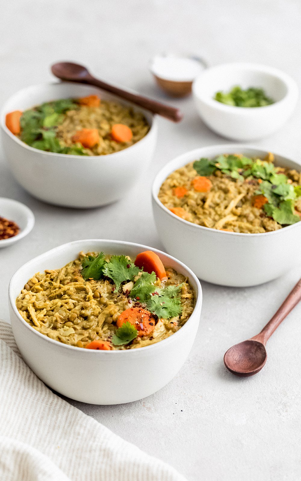 Sopa sabrosa de pollo y lentejas hecha con leche de coco cremosa y rellena con las bondades de la cúrcuma, el jengibre, el ajo y el comino. ¡Esta sopa cremosa de pollo y lentejas con coco dorado está repleta de proteínas y es el almuerzo o la cena perfectos y abundantes! Tres tazones de sopa de pollo y lentejas con cuchara de madera