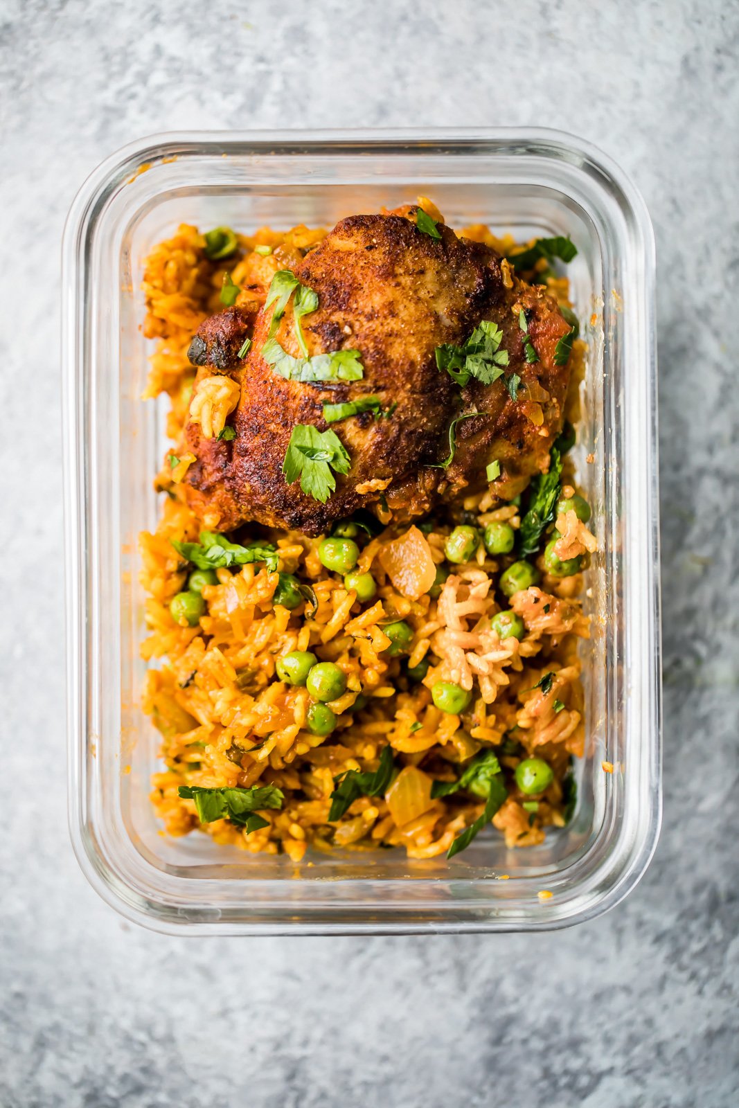 El famoso pollo y arroz puertorriqueño de mi mamá, también conocido como arroz con pollo, está hecho con sofrito de pollo sazonado con adobo casero y arroz sabroso en una sola sartén. Es un plato reconfortante, fácil de preparar y la comida nutritiva perfecta entre semana. ¡Haré esta receta una y otra vez! arroz con pollo en un recipiente de preparación de comida adornado con cilantro