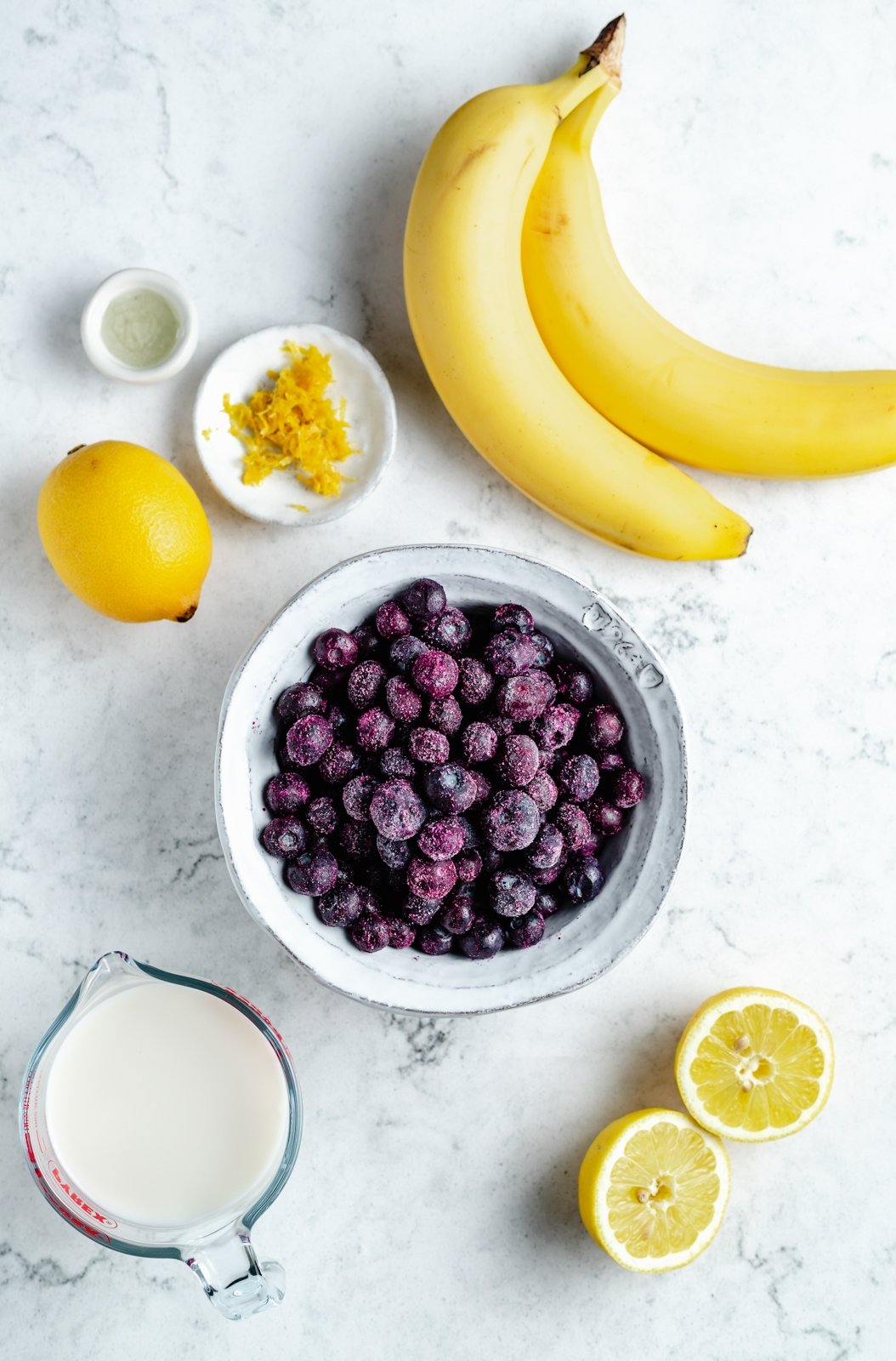 Magnífico batido de limón y arándanos hecho con jugo de limón fresco y jugosos arándanos para una explosión de sabor. Este sencillo batido vegano de limón y arándanos está endulzado naturalmente con fruta y hecho con solo cinco ingredientes simples. Opción de agregar su fortificante nutricional favorito para el perfecto desayuno o merienda! Ingredientes para batido de limón y arándanos en superficie de mármol