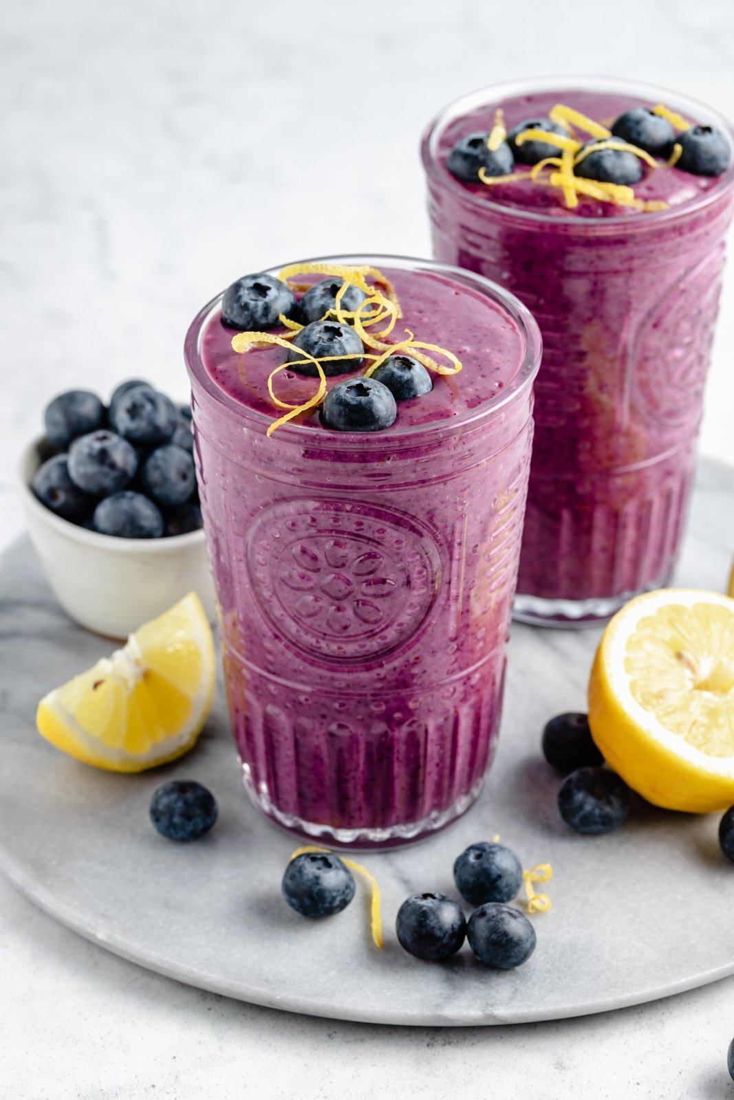 Un delicioso batido de limón y arándanos con sabores brillantes de jugo de limón fresco y jugosos arándanos. Este sencillo batido vegano de limón y arándanos se endulza de forma natural con fruta y se prepara con solo cinco ingredientes simples. ¡Opción de agregar su refuerzo nutricional favorito para el desayuno o merienda perfecto! Dos vasos de batido de limón y arándanos en una bandeja