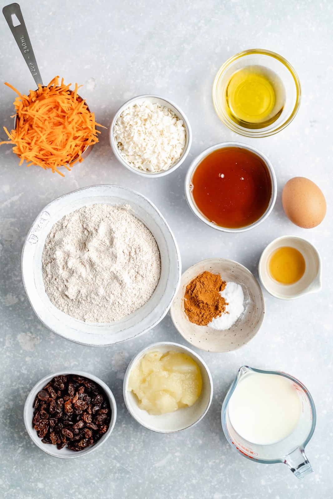 Muffins de pastel de zanahoria saludables rellenos de zanahorias, coco, pasas y nueces. Estos muffins de pastel de zanahoria deliciosamente húmedos están endulzados de forma natural con jarabe de arce puro y compota de manzana. Cubiertos con un glaseado de queso crema ligero, sirva como un refrigerio delicioso o como un saludable ¡merienda! Ingredientes para muffins de pastel de zanahoria en tablero gris