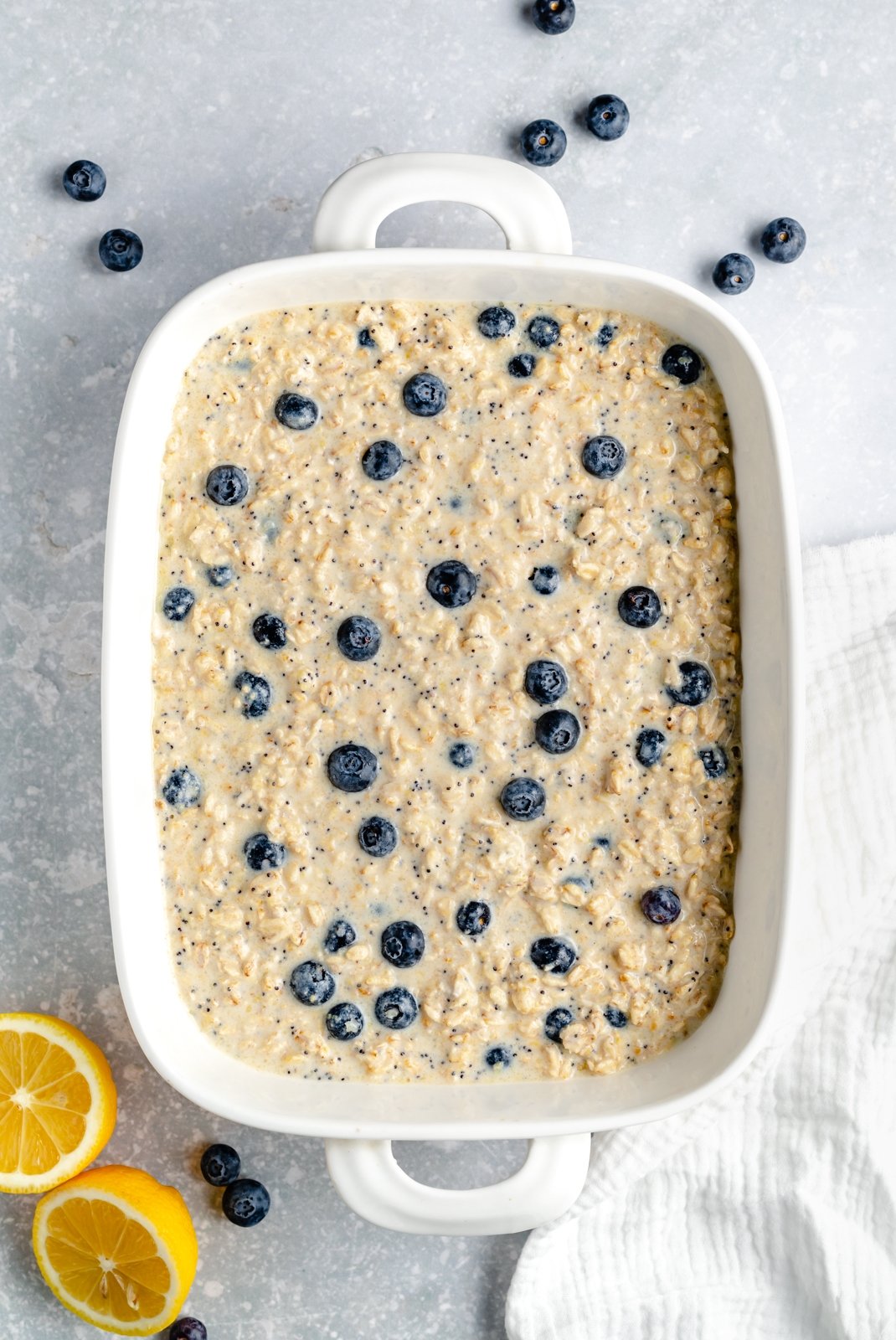 Hermosa avena horneada con limón y arándanos hecha con jugo de limón fresco y cáscara y endulzada naturalmente con jarabe de arce puro. Esta avena horneada con limón no contiene gluten ni lácteos y contiene arándanos frescos y semillas de amapola en cada bocado. Rociada con cobertura casera de yogur de limón es ¡el regalo perfecto para el desayuno! Avena horneada con limón y arándanos sin hornear en una bandeja para hornear