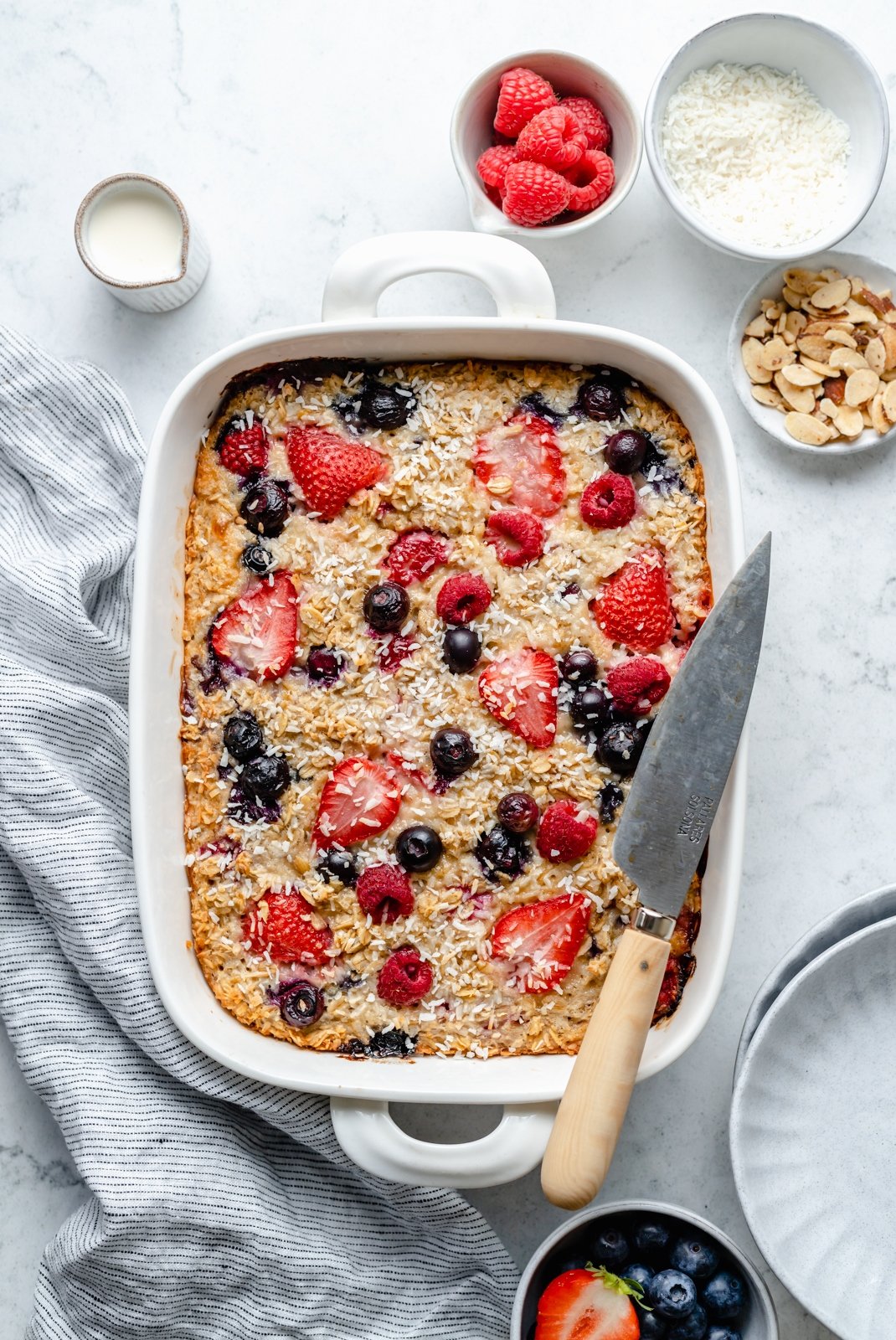 Maravillosa avena horneada con crema y bayas triples hecha con fresas, arándanos y frambuesas y endulzada naturalmente con un toque de jarabe de arce puro. ¡Esta receta fácil de avena horneada con bayas presenta grasas saludables en leche de coco cremosa y es la manera perfecta de usar bayas frescas! Avena de bayas horneada en una bandeja para hornear con un cuchillo