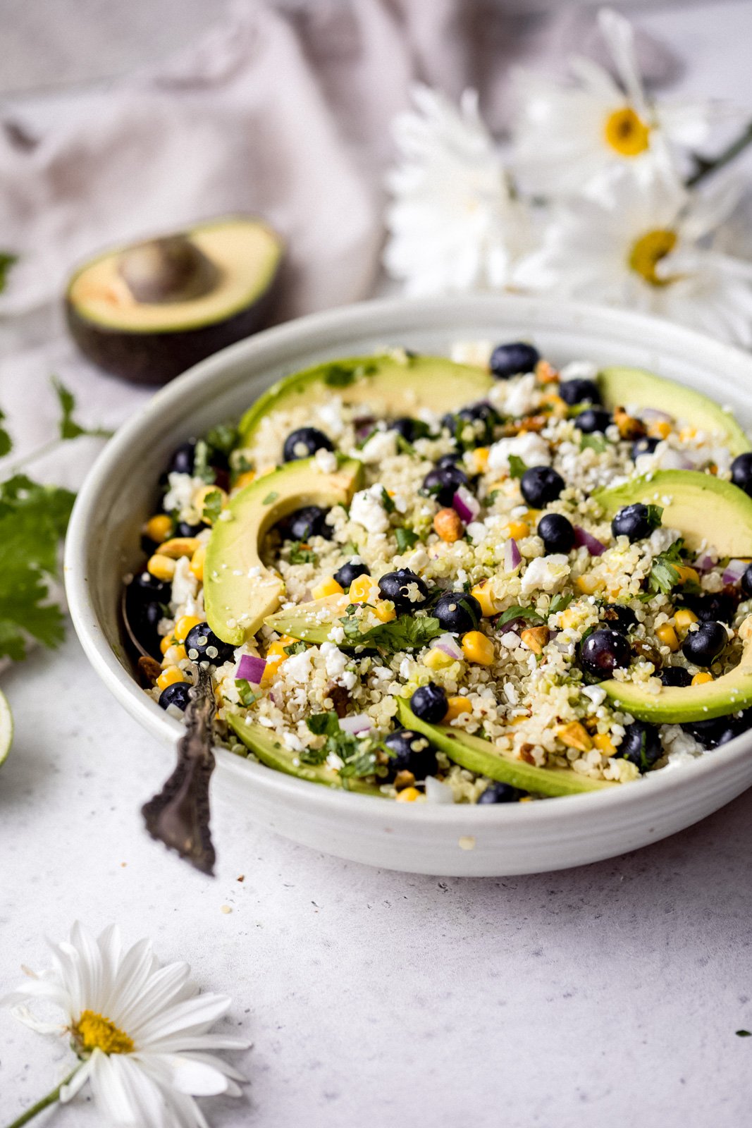 Magnífica ensalada de aguacate, arándanos y quinua con maíz fresco, cebolla roja, queso feta picante, pistachos picados y mezclada con un delicioso aderezo de cilantro y lima. Esta deliciosa ensalada vegana de quinua es perfecta para almuerzos de verano, fiestas y comidas al aire libre. Ensalada De Aguacate Y Arándanos Con Una Cuchara En Un Tazón