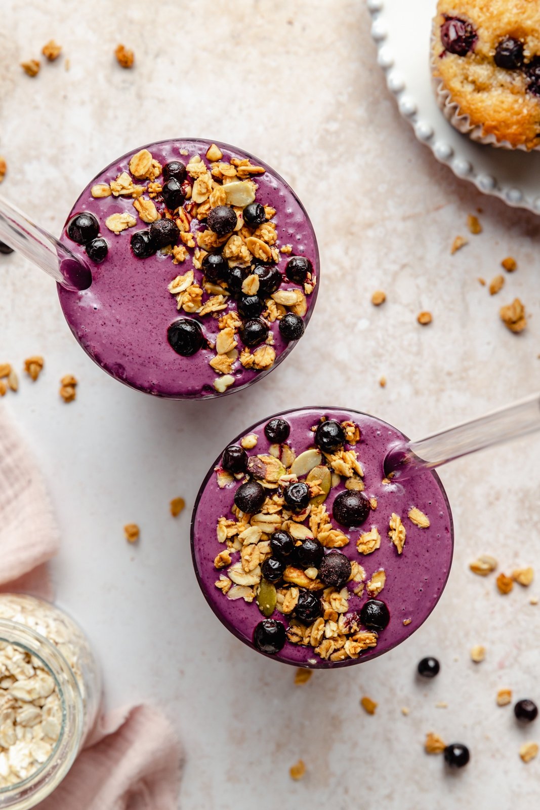 ¡Un batido de arándanos increíble hecho con arándanos silvestres que sabe igual que los muffins de arándanos recién horneados! Este licuado de muffin de arándanos rico en nutrientes es naturalmente dulce sin plátanos, repleto de proteínas de yogur griego y lleno de sabor a muffin gracias a la avena y el lino. ¡Este delicioso desayuno o merienda incluye complementos de sabor opcionales! Vista superior de dos batidos de arándanos cubiertos con avena