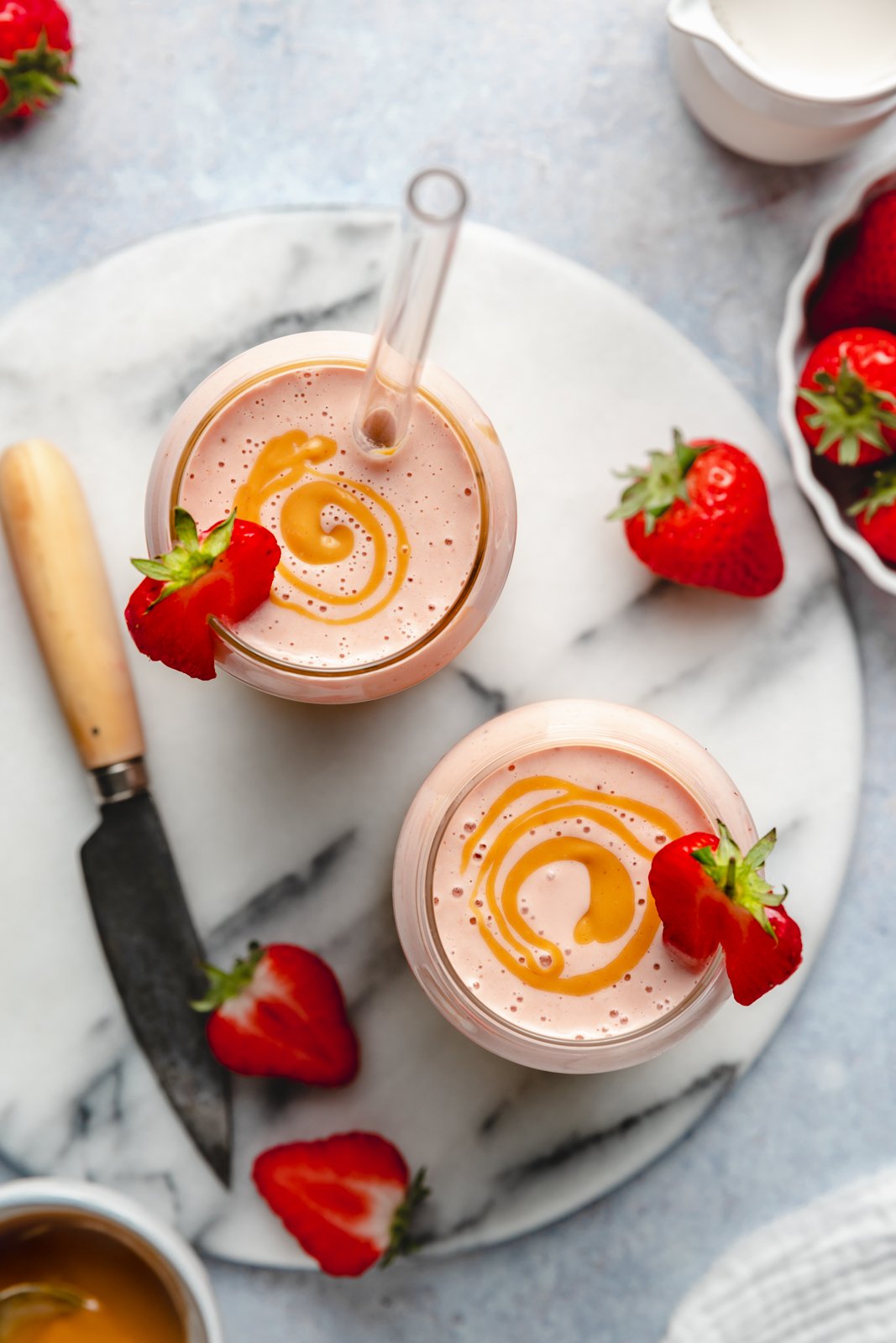 El batido de plátano, fresa y mantequilla de maní más cremoso que te deja desnudo y delicioso. ¡Solo necesitas 5 minutos y 4 ingredientes simples para hacer este increíble batido con un sabor nostálgico de plátano, mantequilla de maní y mermelada! Este batido de mantequilla de maní, plátano y fresa es vegano y está repleto de grasas saludables y proteínas, lo que lo convierte en el desayuno o merienda perfecto. Vista de arriba hacia abajo de dos batidos de plátano, fresa y mantequilla de maní en vasos