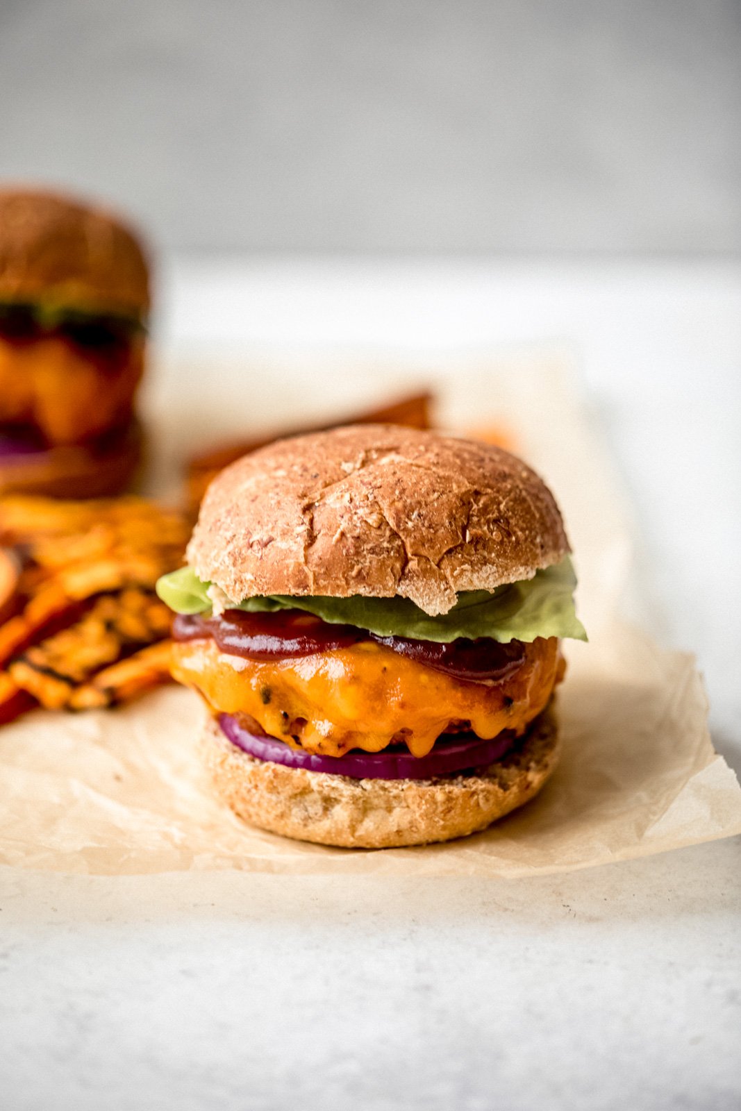 Muy buenas hamburguesas de pollo a la barbacoa y batatas ralladas para mantenerlas jugosas. Cubra estas increíbles hamburguesas de pollo con camote con salsa BBQ dulce y picante, queso cheddar fuerte, cebolla roja y lechuga para obtener la mejor receta saludable de hamburguesa de pollo a la parrilla, ¡perfecta para el verano! Hamburguesas de pollo con batata con queso, salsa BBQ y lechuga en papel pergamino y servidas con papas fritas con batata