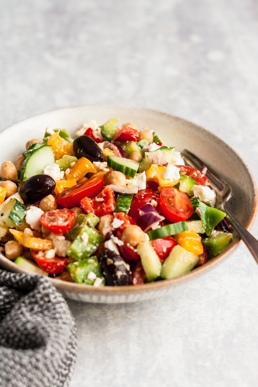 Esta receta fácil de ensalada griega picada toma menos de 20 minutos y está repleta de sabores mediterráneos, vegetales coloridos y proteína vegetal de garbanzos. Te encantará esta ensalada griega de garbanzos porque es rápida y fácil para el almuerzo o para tomar ¡a una fiesta! Ensalada griega picada con garbanzos en un bol con un tenedor