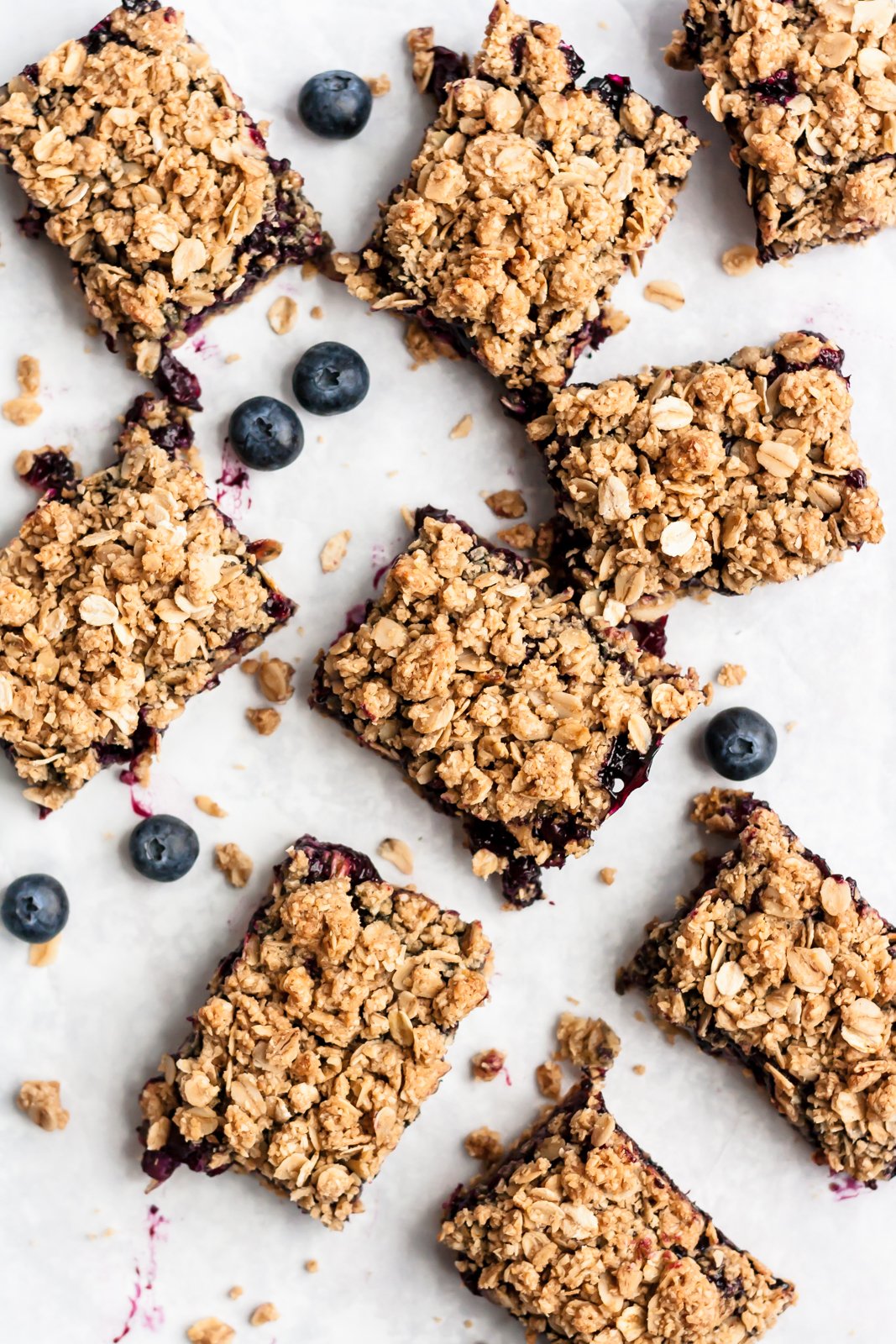 Barritas de pastel de arándanos muy dulces y saludables cubiertas con deliciosas migajas de avena. ¡Estas sencillas barritas de arándanos veganas y sin gluten saben igual que la tarta de arándanos y serán tus nuevas favoritas de primavera y verano! Tiras de tarta de arándanos esparcidas sobre una superficie blanca rodeada de arándanos