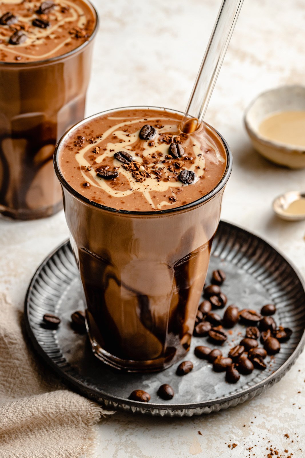 Espeso y delicioso batido de café con tahini y dátiles, elaborado con cremosa leche de coco y endulzado naturalmente con dátiles. ¡Esta receta fácil de batido de café con dátiles tiene un toque de rico sabor a chocolate del cacao en polvo e incluso vegetales furtivos! Te encantará este batido para un excelente desayuno o un refrigerio por la tarde. Tahini Coffee Date Shake y granos de café en un plato