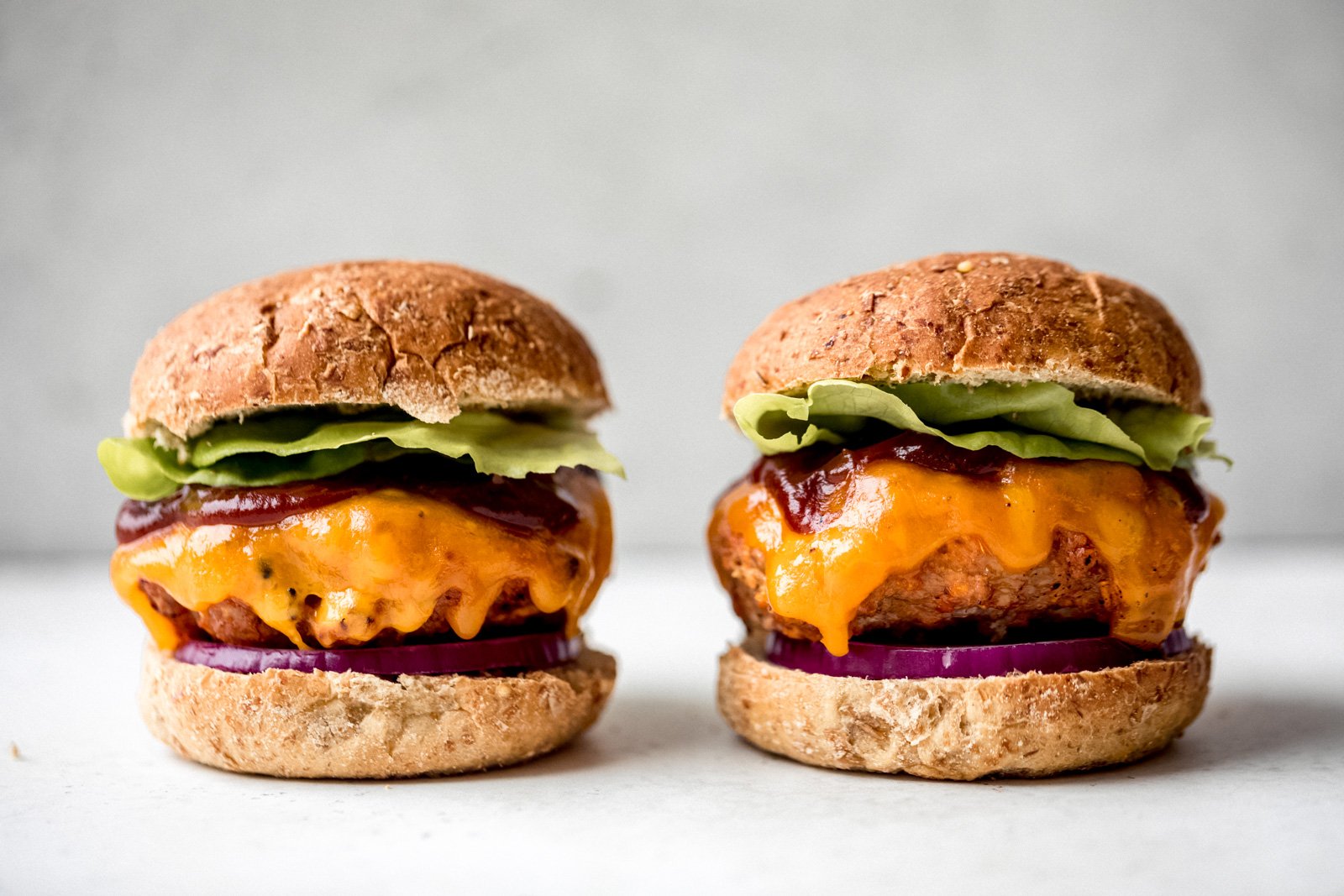 Excelentes hamburguesas de pollo con batatas ralladas para mantenerlas jugosas. Cubierto con una salsa BBQ dulce y ácida, queso cheddar fuerte, cebolla morada y lechuga, ¡esta es la receta de hamburguesa de pollo a la parrilla saludable y perfecta para el verano! Dos hamburguesas de pollo BBQ adyacentes cubiertas con queso, salsa BBQ y lechuga