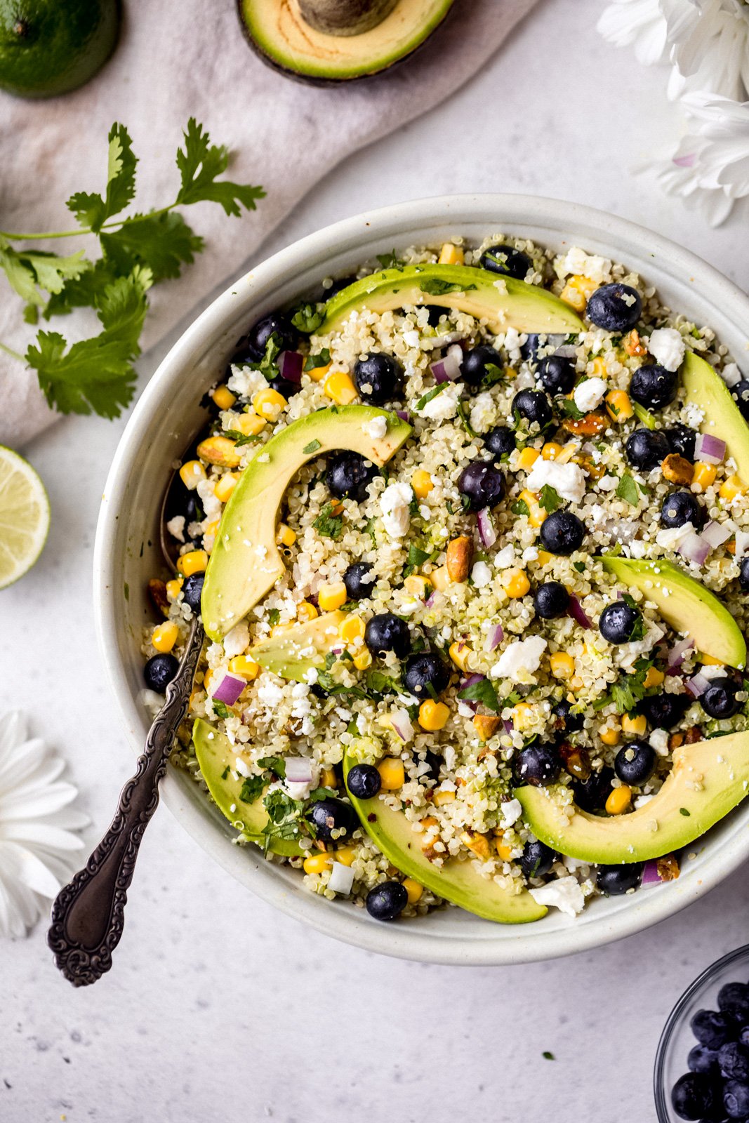 Magnífica ensalada de aguacate, arándanos y quinua con maíz fresco, cebolla roja, queso feta picante, pistachos picados y mezclada con un delicioso aderezo de cilantro y lima. Esta deliciosa ensalada vegana de quinua es perfecta para almuerzos de verano, fiestas y comidas al aire libre. Ensalada de aguacate, arándanos y quinua en un tazón con una cuchara a la izquierda
