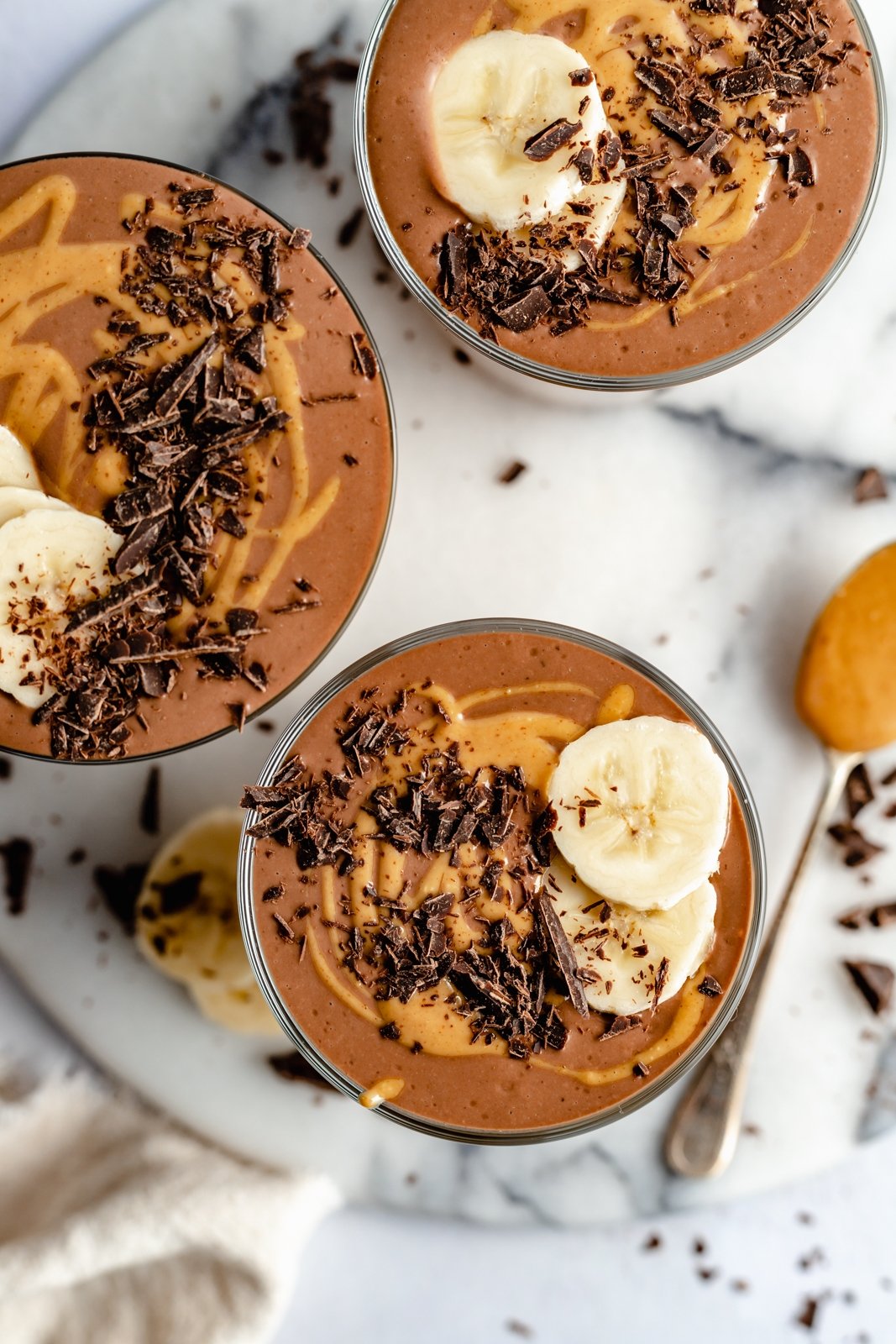 ¡Un delicioso batido cremoso de chocolate con mantequilla de maní y plátano que realmente sabe como el batido de Wendy's! Esta saludable receta de batido de chocolate y plátano está hecha con solo cinco ingredientes y es fácil de usar con su refuerzo nutricional favorito Personalizado. Perfecto para el desayuno, la merienda o incluso el postre ! Vista de arriba hacia abajo de tres batidos saludables de chocolate y plátano en vasos