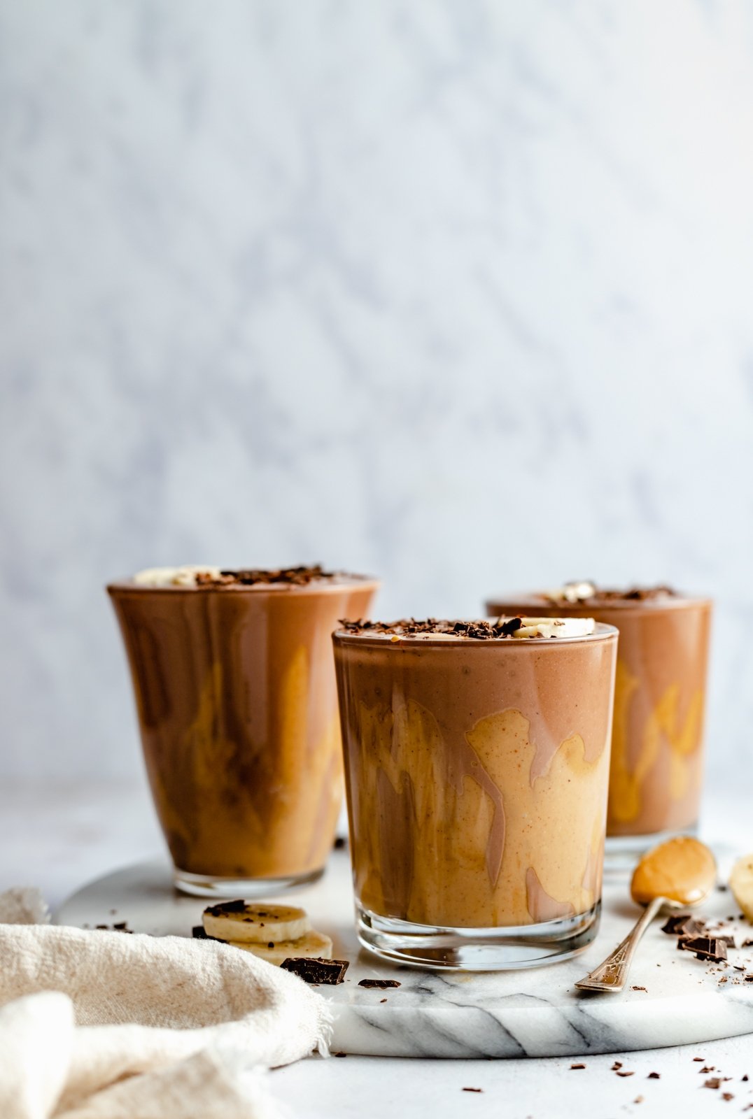¡Delicioso batido cremoso de chocolate con mantequilla de maní y plátano que sabe como el batido de Wendy's! Esta receta saludable de licuado de plátano y chocolate está hecha con solo cinco ingredientes y es fácil de personalizar con su potenciador nutricional favorito. ¡Perfecto para el desayuno, la merienda o incluso el postre! Tres batidos de plátano con mantequilla de maní y chocolate en una placa de mármol