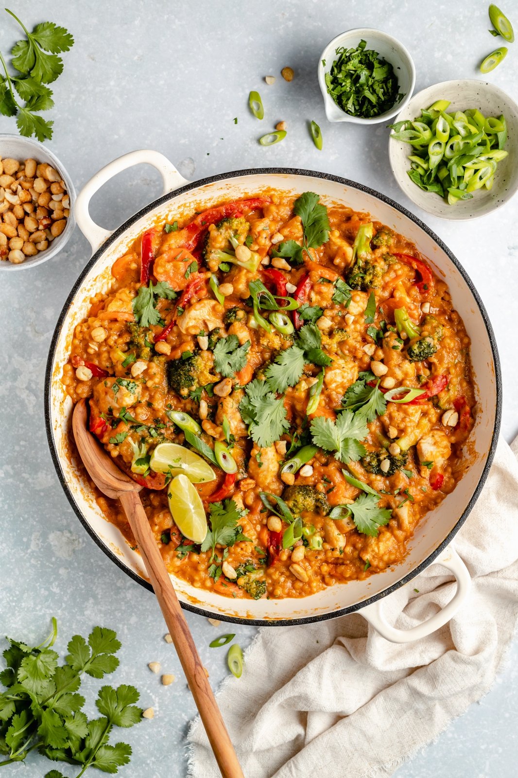 Delicioso cuscús de pollo tailandés en una sola sartén con cacahuates llenos de proteínas y vegetales coloridos. Esta acogedora receta de cuscús de pollo está infundida con sabores tailandeses gracias a una rica salsa cremosa de maní hecha con leche de coco, jengibre fresco, salsa de soya, ajo y ¡Opciones de mantequilla de maní para condimentar las cosas si te gustan las especias! src= Cuscús de pollo tailandés en una olla con madera cuchara con cilantro