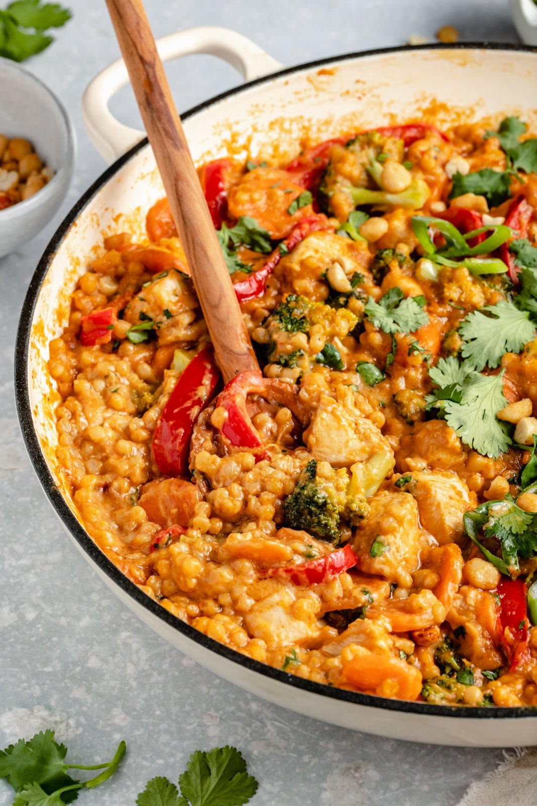 Delicioso cuscús de pollo tailandés en una sola sartén con cacahuates llenos de proteínas y vegetales coloridos. Esta acogedora receta de cuscús de pollo está infundida con sabores tailandeses gracias a una rica salsa cremosa de maní hecha con leche de coco, jengibre fresco, salsa de soya, ajo y ¡Opciones de mantequilla de maní para condimentar las cosas si te gustan las especias! src= Cuscús tailandés con pollo y maní en una sartén con la cuchara de madera