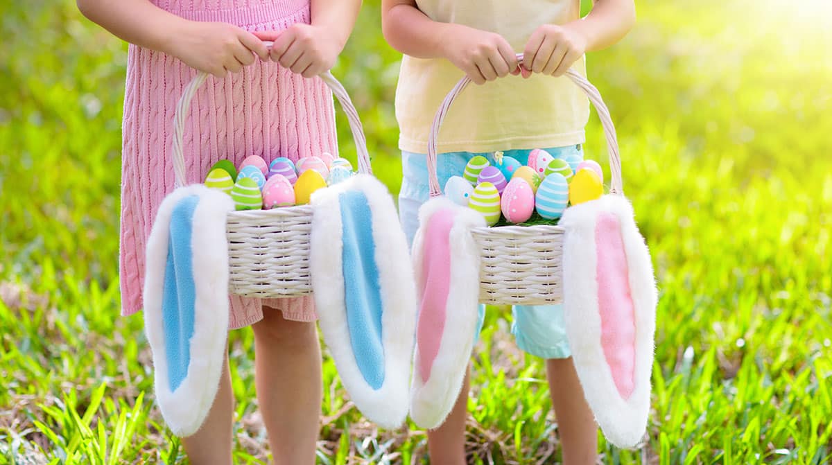 Niños con una canasta de huevos y orejas de conejo en una búsqueda de huevos de Pascua en un soleado jardín de primavera.  El niño y la niña buscan dulces coloridos y huevos de chocolate con una canasta de conejo.  Los niños celebran la Pascua.