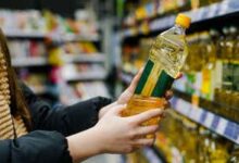 2HKYX4G Mujer elige aceite de girasol en supermercado. Primer plano de una mano sosteniendo una botella de aceite en la tienda.