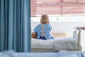 Vista trasera de una niña enferma con un juguete suave sentado en la cama. Una paciente infantil está descansando en la sala de una clínica. Llevaba una bata de hospital.