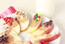 Snacks de Manzana - Saludables, Equilibrados, Sencillos 13 Postre de manzana con rodajas de manzana, mantequilla de maní y varios ingredientes en un plato blanco
