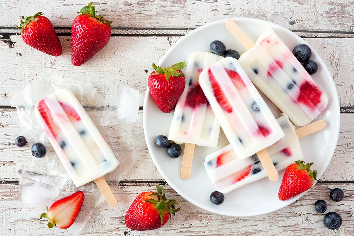 Paletas de yogur de fresa y arándanos saludables en un plato, sobre un fondo de madera blanca, con vistas a una escena de mesa de verano
