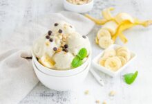 banana ice cream in a bowl with peanuts and chocolate on a wooden background with sliced bananas in the background