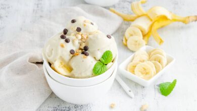 banana ice cream in a bowl with peanuts and chocolate on a wooden background with sliced bananas in the background