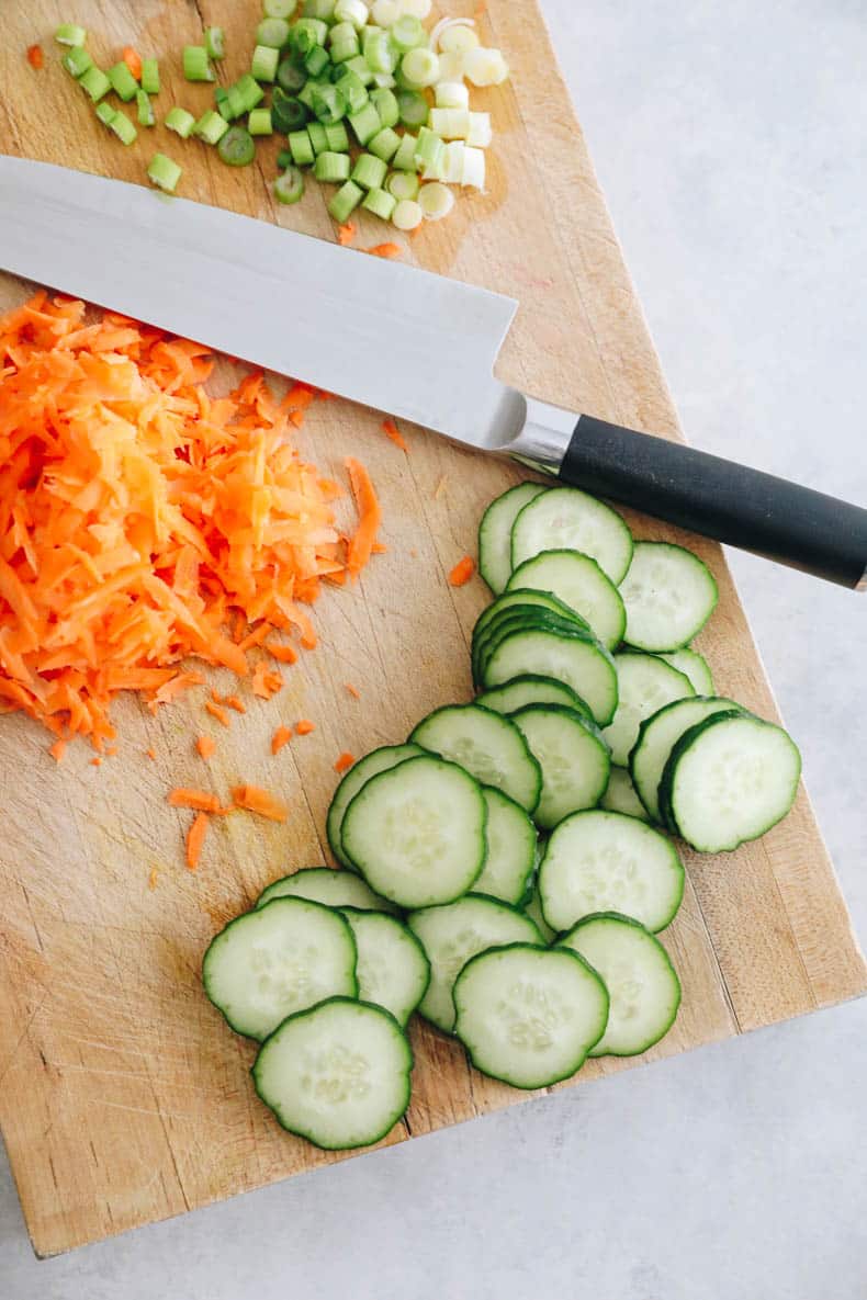 Tabla de cortar con pepinos en rodajas, zanahorias ralladas y cebollas verdes picadas.