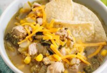 Green chili in a white bowl with tortilla chips and shredded cheddar cheese.